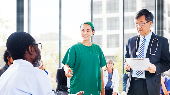 A male doctor and a female nurse speaking to a small group of diverse people sitting in a circle.